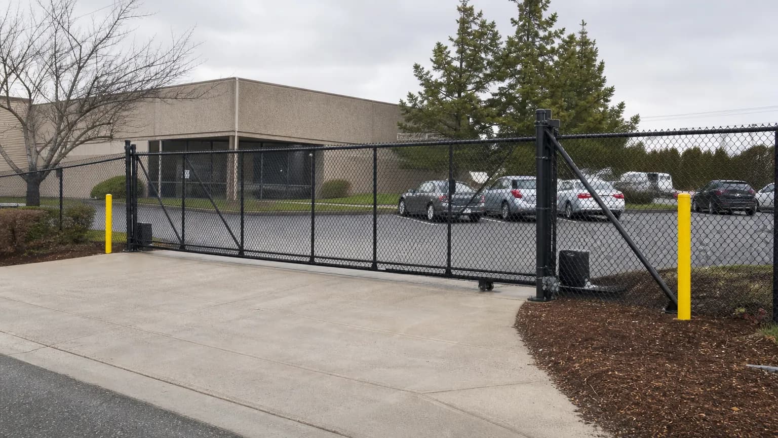 Tall black vinyl-coated chain link security fence with a rolling gate around a small business parking lot under an overcast sky