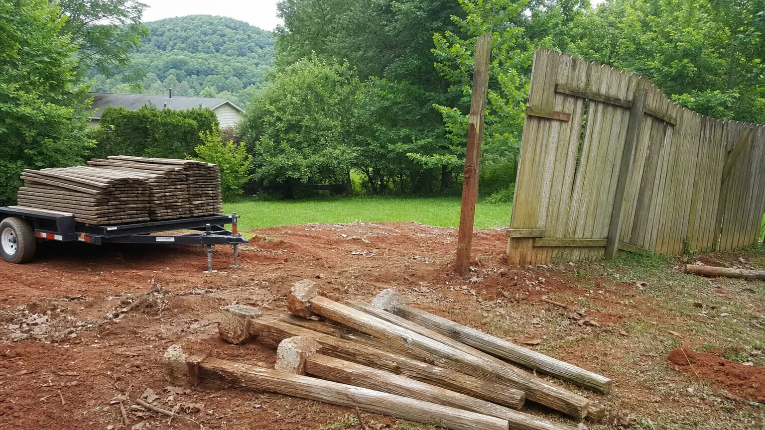 Old leaning wood privacy fence half torn down with pickets stacked on a trailer and pulled posts on red clay